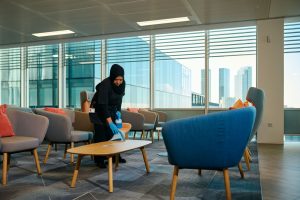 A person is cleaning a modern office lounge, wiping a table with a cloth, surrounded by colourful chairs and large windows.