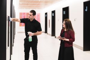 A man is cleaning a wall with spray while a woman observes, holding a tablet in a bright hallway.
