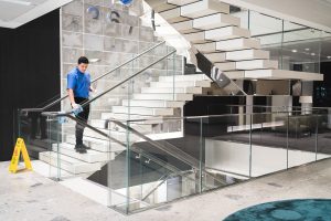 A person in blue cleans a modern glass staircase with a cloth, ensuring a spotless finish. A caution sign is visible nearby.